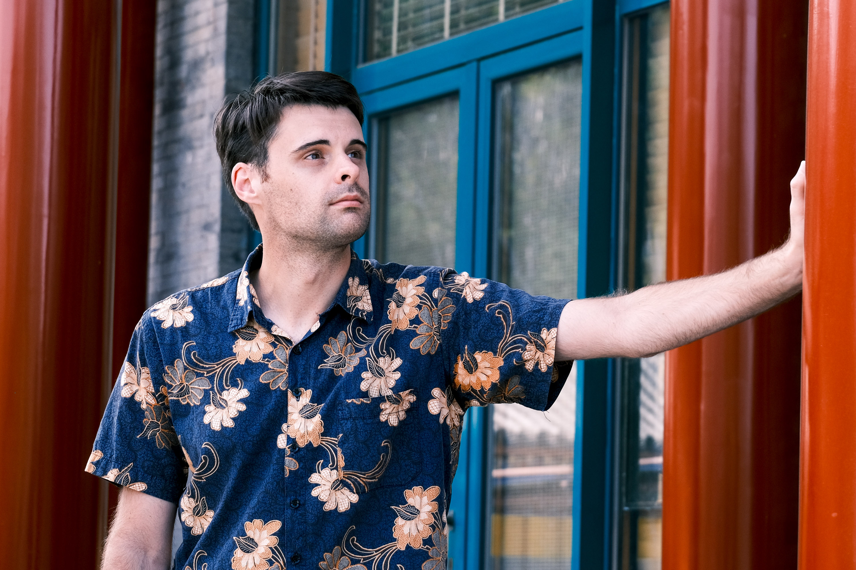 A photograph of a thirty-year-old man standing in front of a grey brick building, leaning on a bright red pole with one hand, while gazing into the sky at nothing in particular. He is posing like this because his lovely wife, who took the photograph, told him to do so. He has handsome features and dark brown hair that he cut himself. He is wearing a dark blue short-sleeved shirt with a floral pattern which he bought in Malaysia.